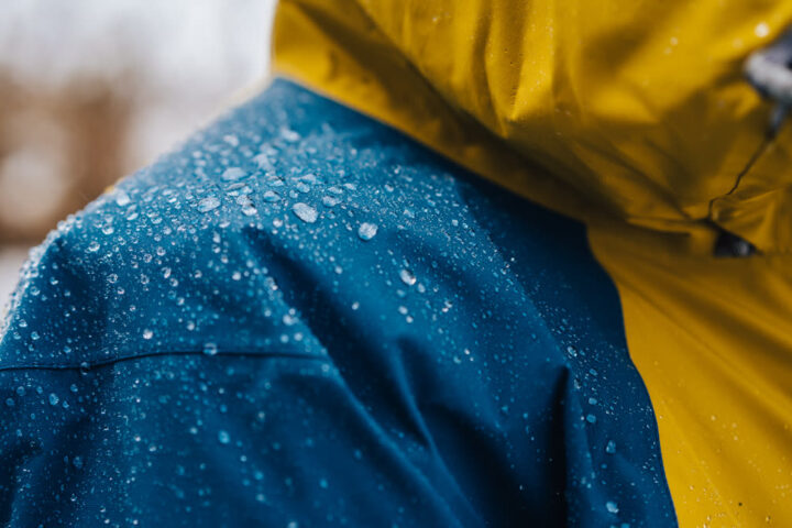 Ilustračný stock: outdoor bunda v daždi, detail membrány, bez čitateľnej značky Gore-Tex ani TNF a bez hero tváre modelu.
