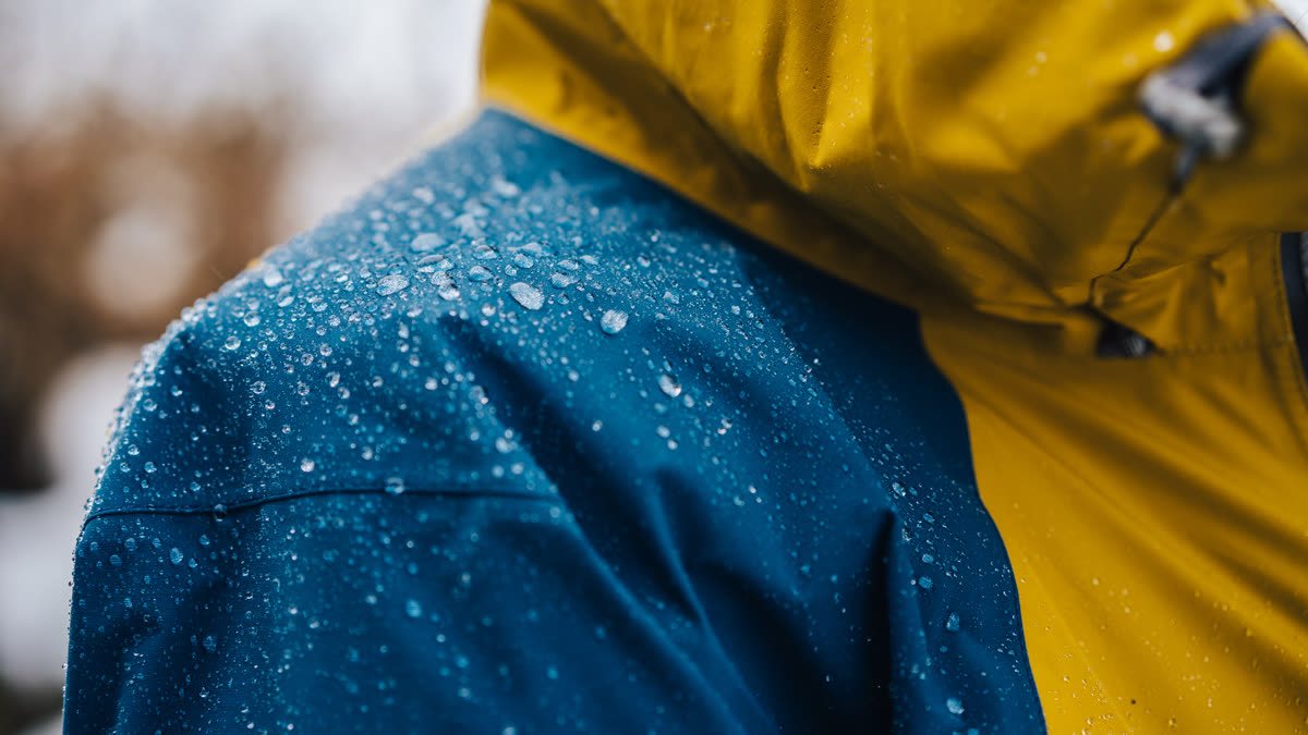 Ilustračný stock: outdoor bunda v daždi, detail membrány, bez čitateľnej značky Gore-Tex ani TNF a bez hero tváre modelu.